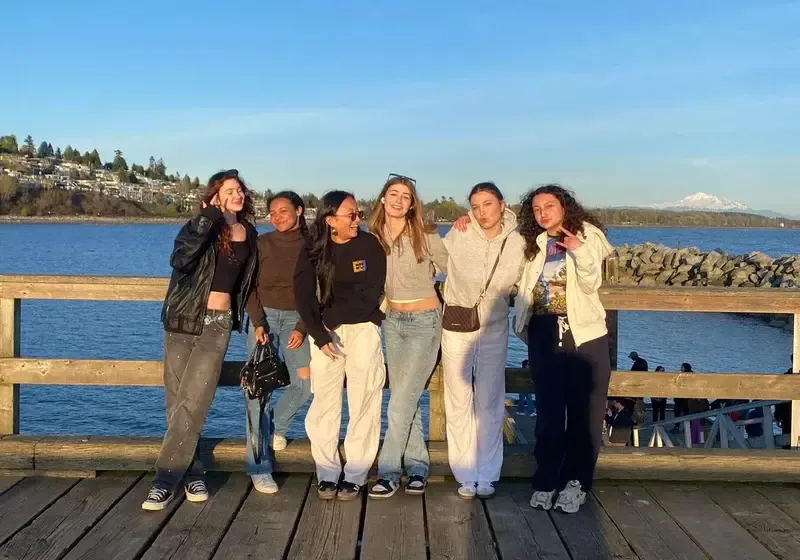 Seis chicas posando en un muelle de madera junto al mar con una montaña nevada al fondo