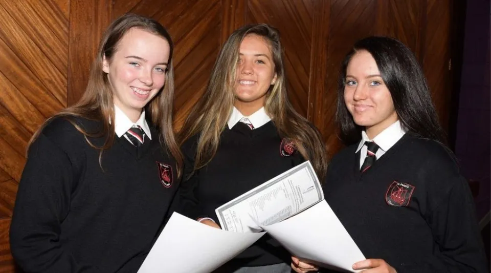 Tre studentesse in divisa scolastica tengono dei fogli e sorridono davanti a un rivestimento in legno nella St. Peter's Community School.