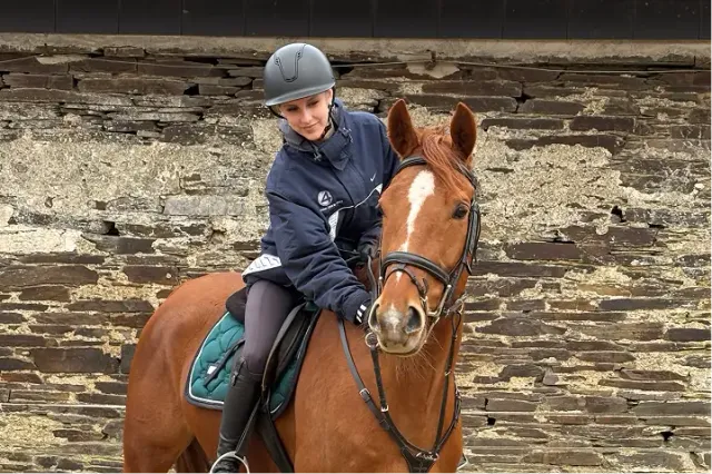 Ragazza con casco a cavallo di un cavallo baio davanti a un muro di pietra