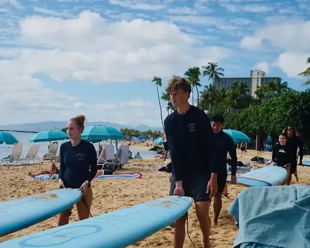 Alumnos de un curso de surf llevando sus tablas por una playa con palmeras y hoteles al fondo