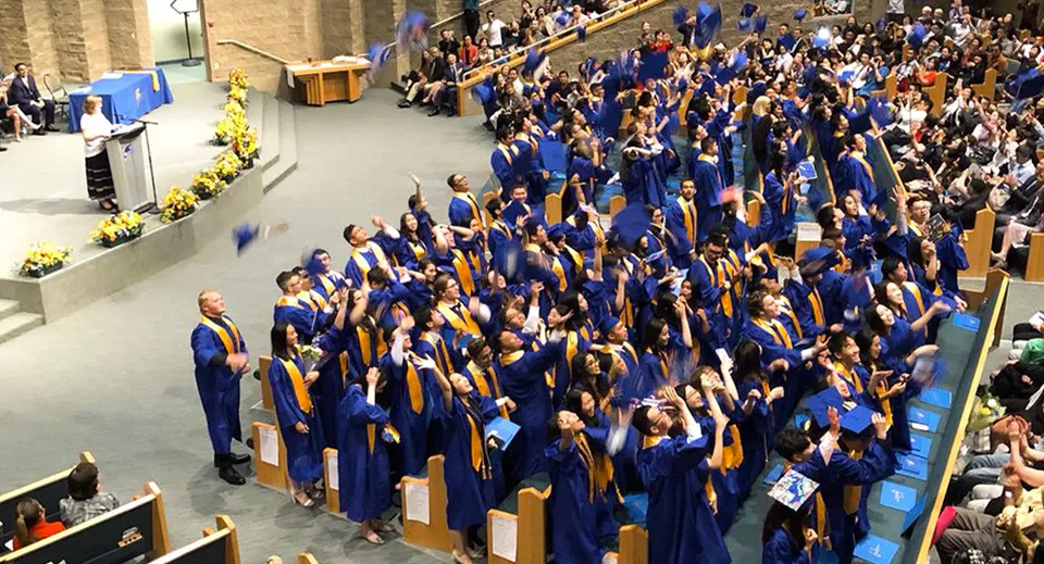 Un grande gruppo di diplomati con toghe blu e dorate si riunisce nell'aula magna della A.R. MacNeill Secondary School durante una cerimonia di diploma.
