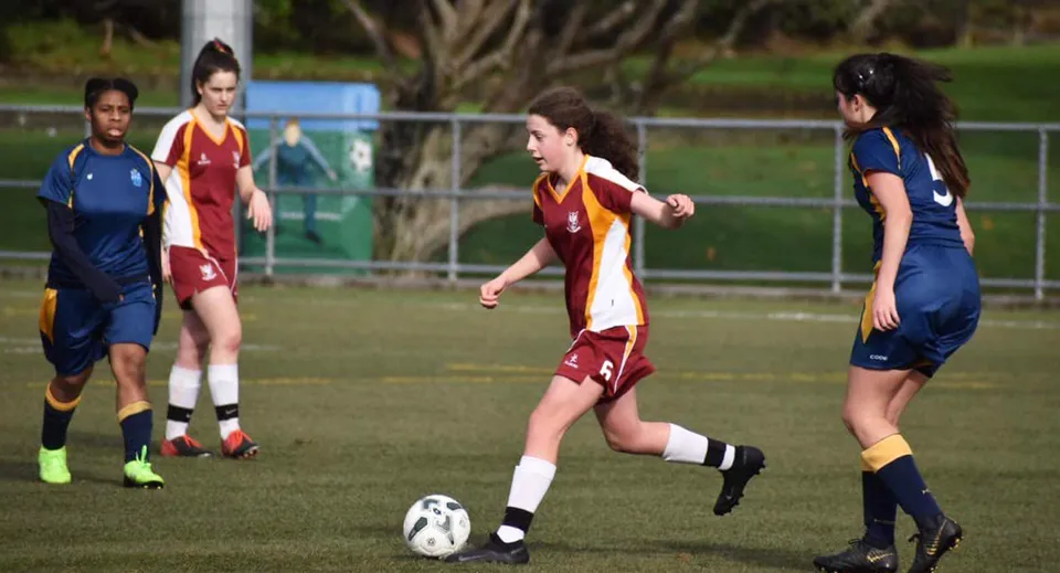 Studentesse del Heretaunga College giocano una partita di calcio su un campo in erba con edifici sullo sfondo.