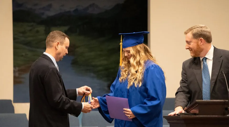 Una studentessa in toga da diploma blu della Maranatha Baptist Academy stringe la mano a un uomo in abito con uno sfondo di montagne e foreste.