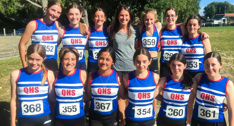 Un gruppo di studentesse della Queens High School indossa uniformi sportive blu e bianche su un campo sportivo.
