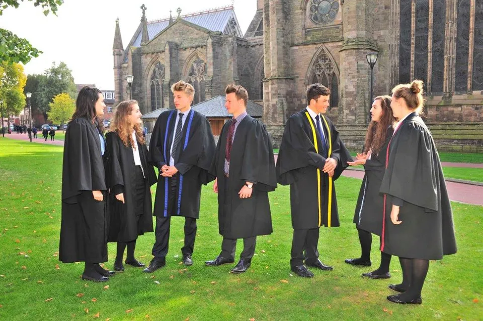 Un gruppo di studenti con la toga di laurea si trova davanti a un grande edificio in pietra nel campus della Hereford Cathedral School.