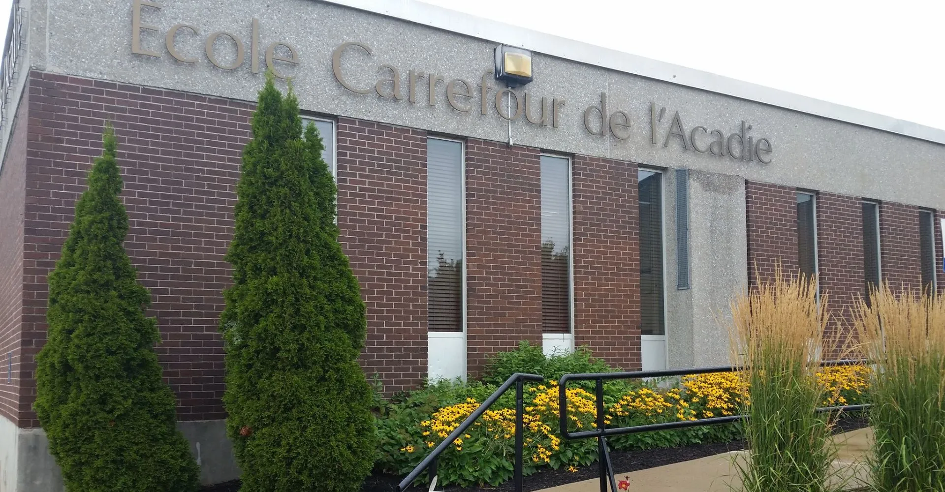 L'edificio in mattoni della scuola Carrefour de l’Acadie è circondato da alti alberi sempreverdi e piante in fiore.