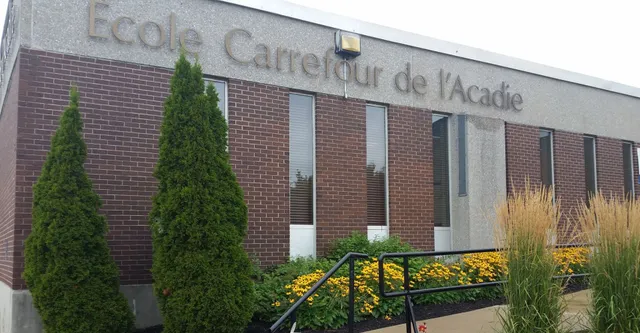 El edificio de ladrillo de la escuela Carrefour de l’Acadie está rodeado de altos árboles perennes y plantas en flor.