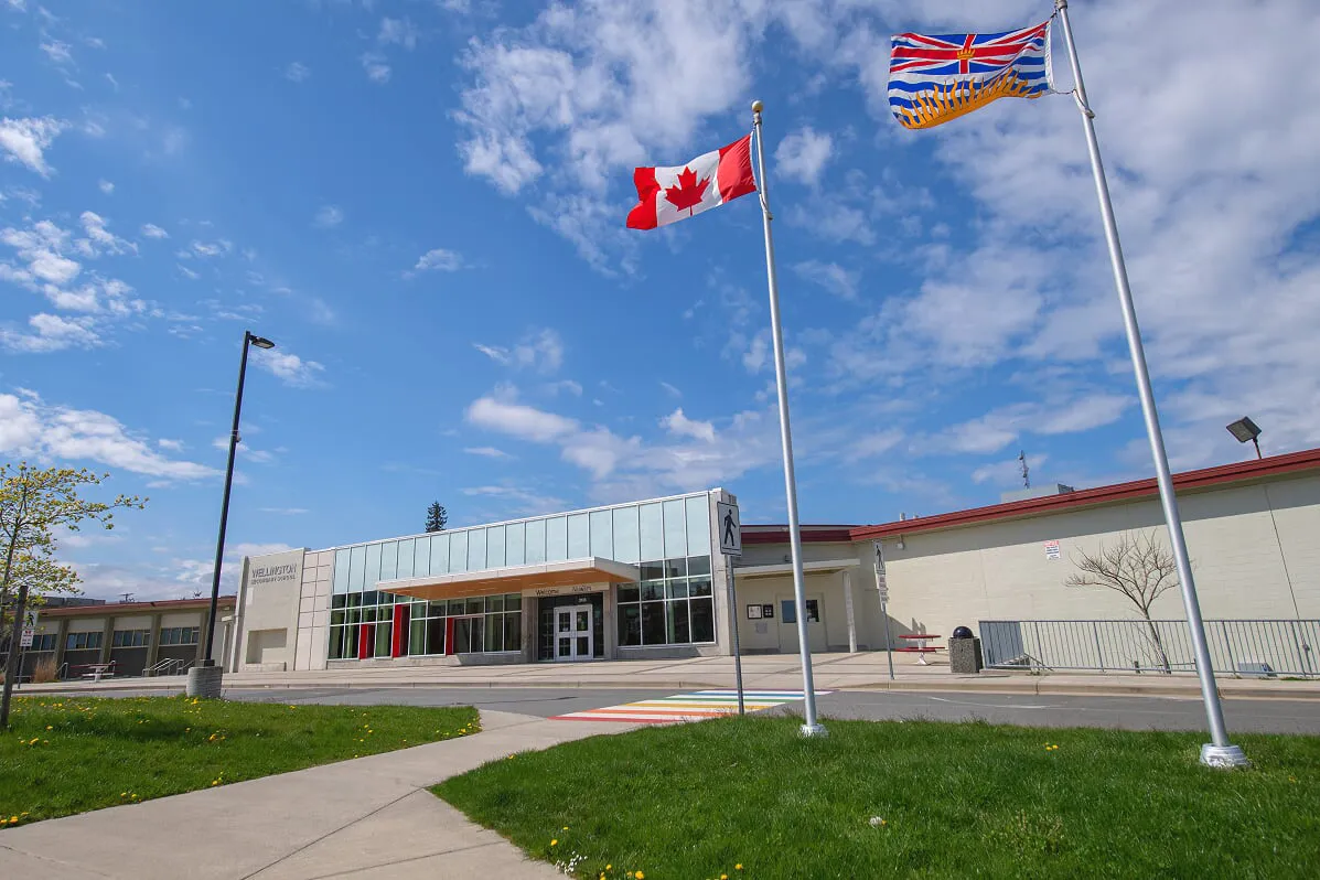 La bandera canadiense ondea sobre el moderno edificio de cristal de Wellington Secondary School ante un cielo azul.