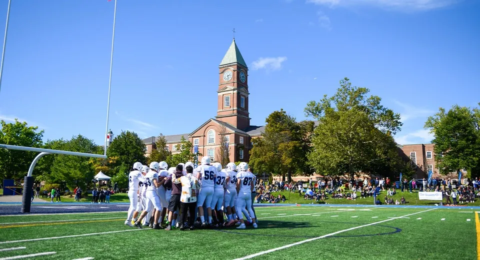 Un gruppo di persone in divisa bianca si raduna su un prato davanti alla torre della chiesa all'interno del campus dell'Upper Canada College.
