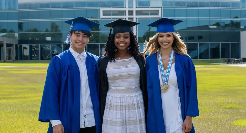 Tre diplomati della Trinity Christian Academy stanno in piedi in toga blu davanti a un edificio moderno con un prato sullo sfondo.