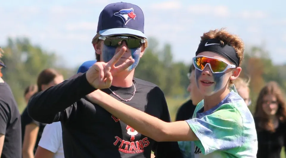 Due persone con cappellini da baseball e occhiali da sole fanno un gesto con le mani davanti a un'area erbosa nel campus della Seaway District High School.