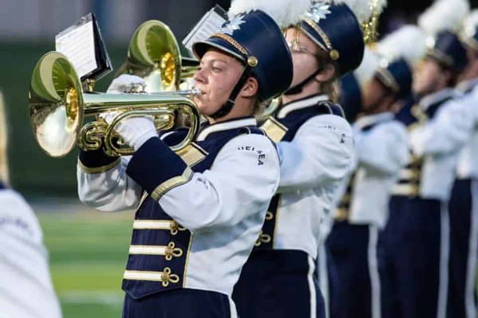 Un membro della Marching Band della Chelsea High School suona uno strumento a fiato in ottone, in uniforme bianca e blu.