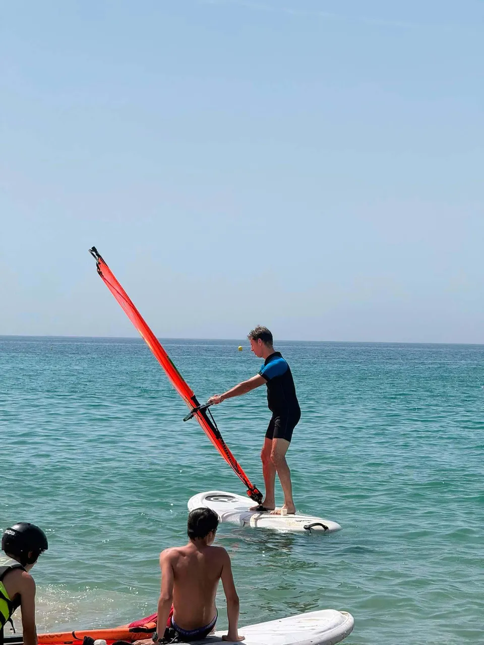 Un uomo su una tavola da windsurf con una vela arancione in mare presso il Novaschool Añoreta College.