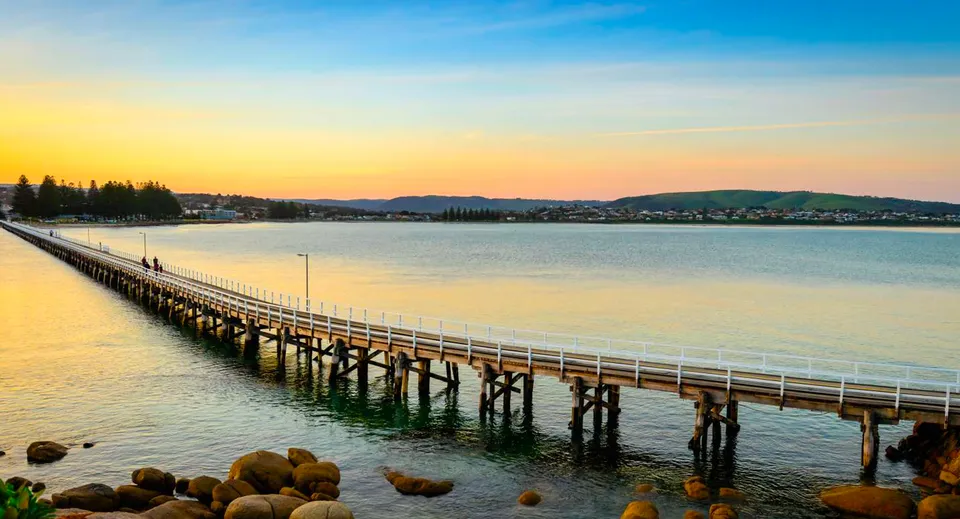 Un lungo pontile in legno si estende nel campus della Victor Harbor High School su un lago tranquillo con colline verdi sullo sfondo.