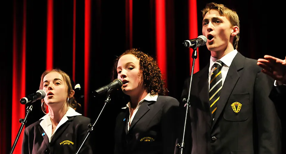 Tre studenti di Awatapu College cantano con i microfoni su un palco davanti a un sipario rosso.