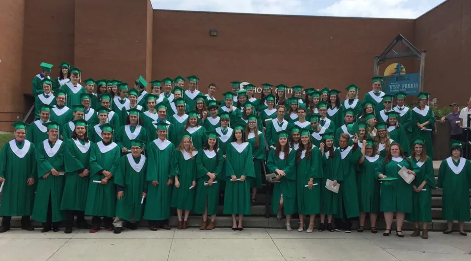 Un gruppo di diplomati in toga e tocco verdi si trova davanti all'edificio in mattoni della West Ferris School con una torre dell'orologio sullo sfondo.