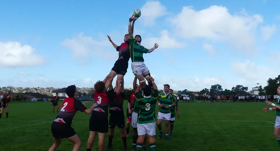 Un gruppo di studenti gioca a rugby sul campo sportivo del Lynfield College sotto un cielo blu con nuvole.