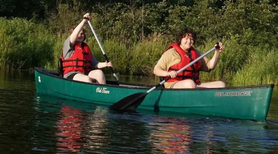 Due studenti della Marvelwood School pagaiano con giubbotti salvagente su una canoa verde in uno specchio d'acqua circondato dalla vegetazione.