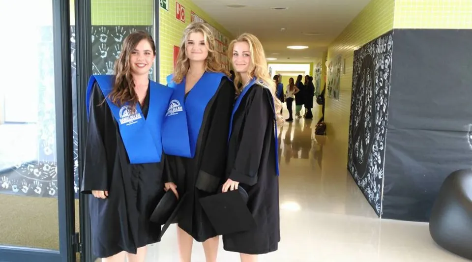 Tre studentesse della Hamelin-Laie International School indossano toghe da diploma blu e nere nel corridoio davanti a una parete-lavagna.