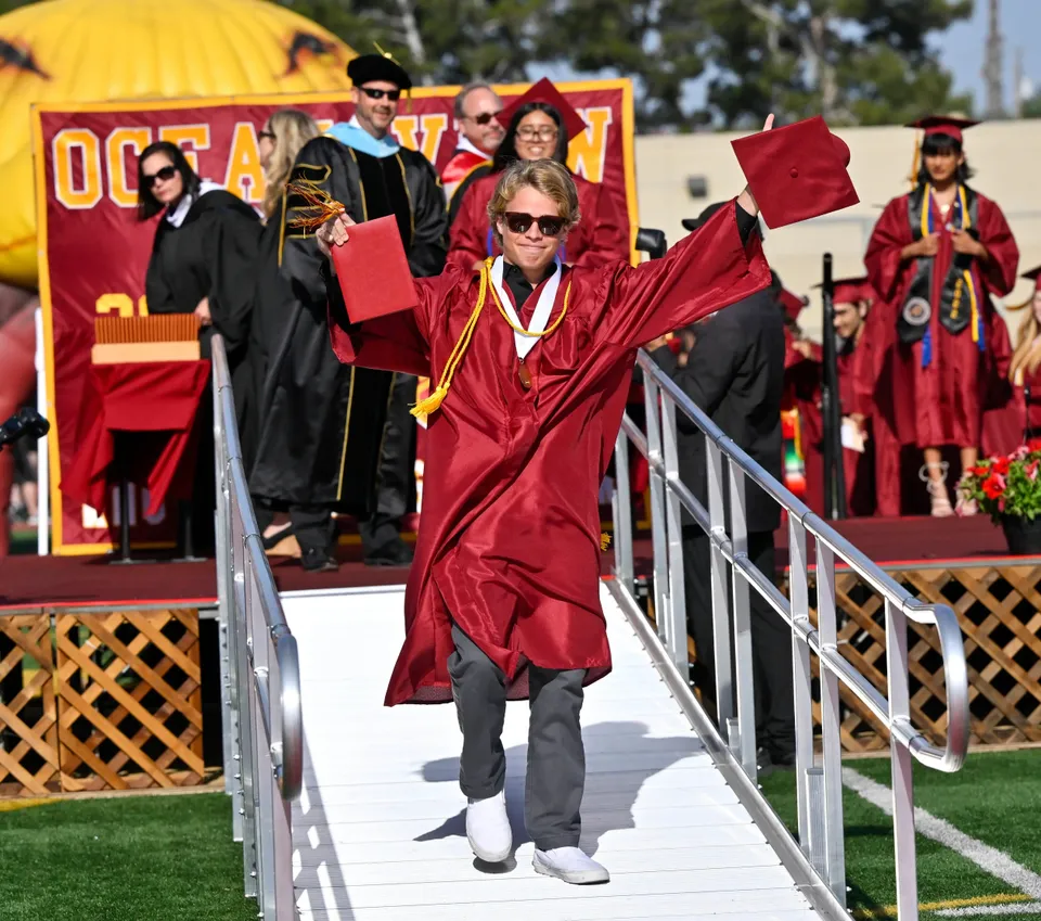 Un diplomato della Ocean View High School in toga rossa e occhiali da sole scende lungo una rampa con il suo diploma.