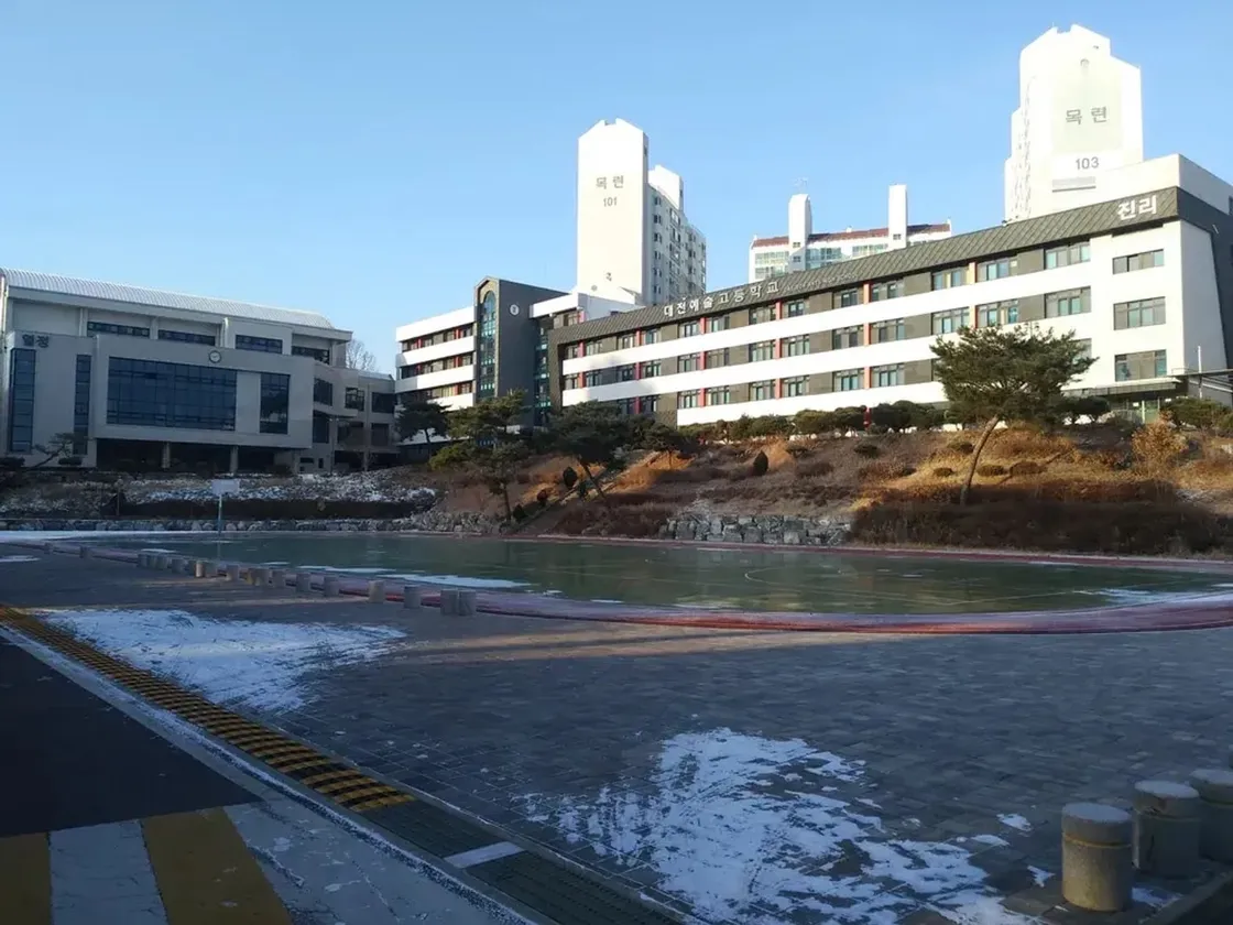 Schulgebäude mit koreanischer Beschriftung und Türmen, davor ein zugefrorener Platz im Winter