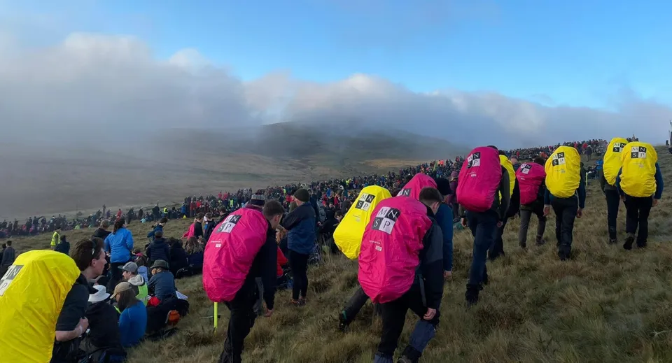 Studenti della Somervale School si trovano su un pendio erboso con abbigliamento da pioggia colorato davanti a montagne nebbiose.