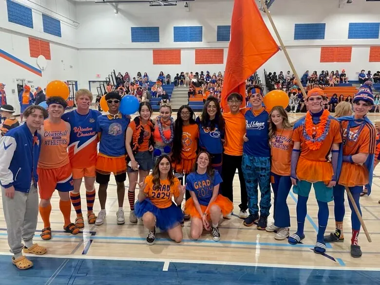 Un gran grupo de estudiantes de la Winston Churchill High School está de pie con sus uniformes naranjas y azules en un gimnasio.