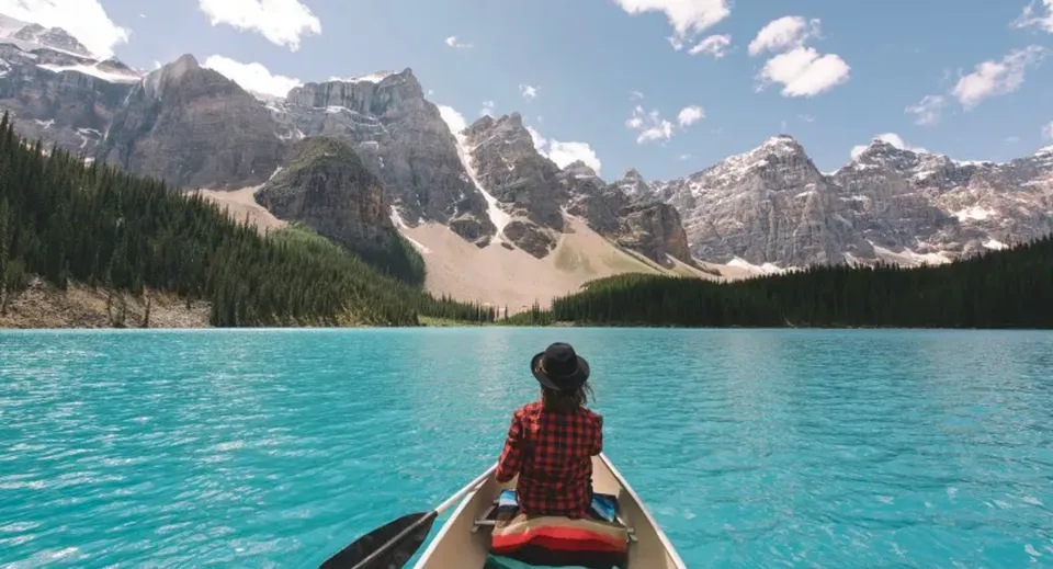 Una persona sta su una piccola barca su un lago calmo davanti a montagne innevate nei pressi della Yale Secondary School.