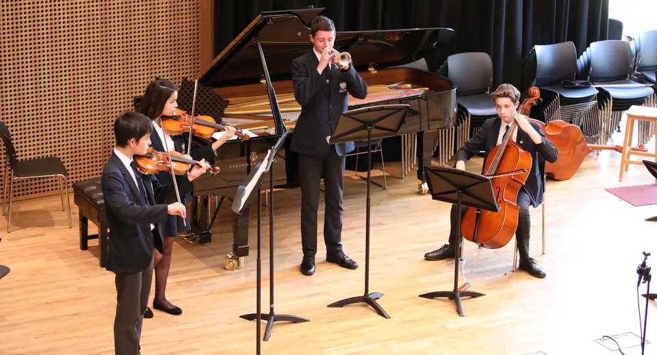 Studenti della Claremont School in abito formale si esibiscono con vari strumenti musicali su un palco davanti a una parete con pannelli di legno.