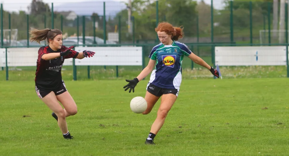 Due studentesse dell'Ursuline College Sligo corrono in abbigliamento sportivo su un campo in erba con un impianto sportivo sullo sfondo.