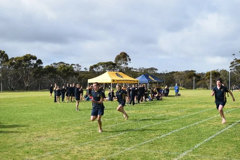 Estudiantes de la Ravensthorpe District High School se reúnen con ropa deportiva en un campo de césped con carpas y árboles al fondo.