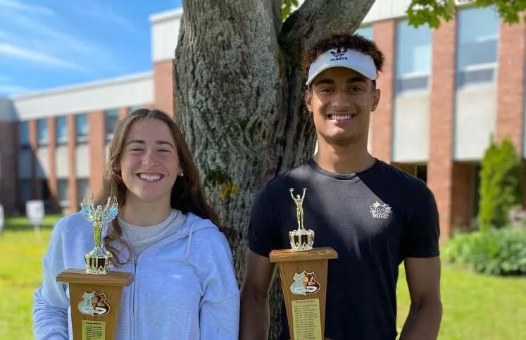 Dos personas están delante de un árbol en el campus de la Colonel Gray High School y sostienen trofeos en la mano.