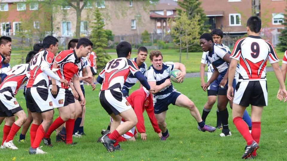 Studenti del Dr. Norman Bethune Collegiate Institute giocano a rugby su un campo sportivo con case sullo sfondo.