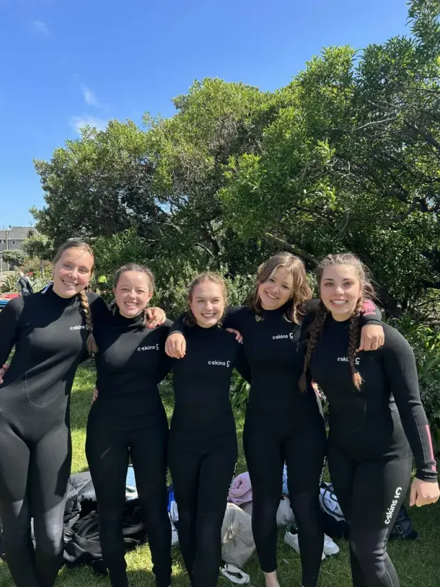 Gruppo di cinque ragazze in muta da surf sorride abbracciato davanti a un albero