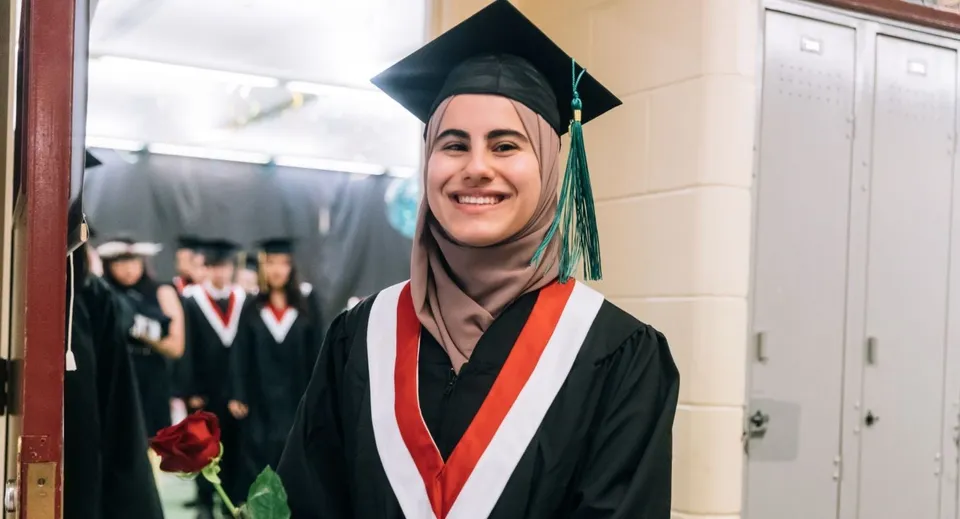 Una diplomata sorridente della Elphinstone Secondary School con hijab, toga e tocco tiene una rosa rossa nel corridoio della scuola.