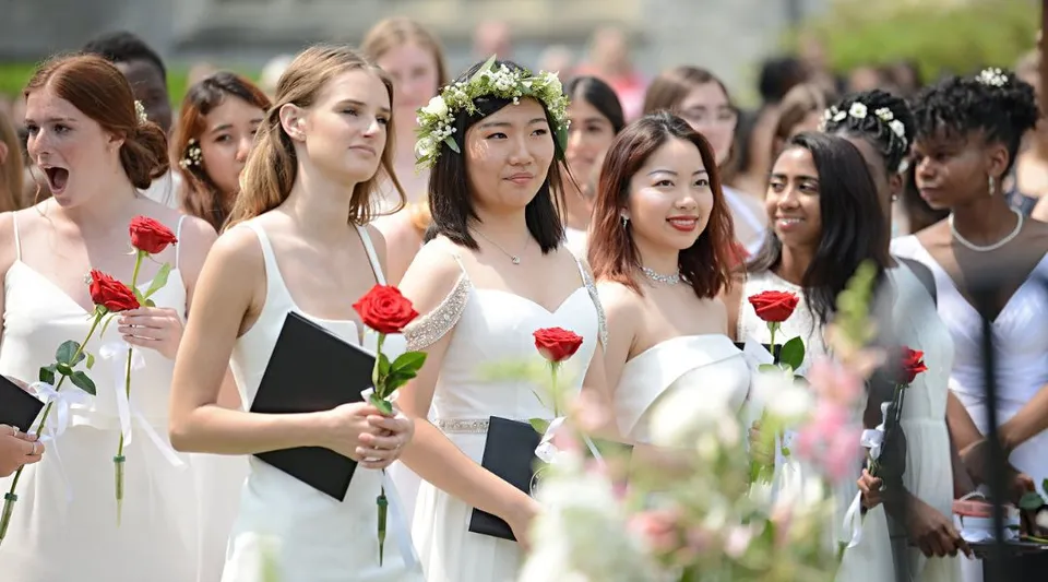 Un gruppo di giovani studentesse della Emma Willard School si trova all'aperto con rose rosse davanti a edifici e aree verdi.