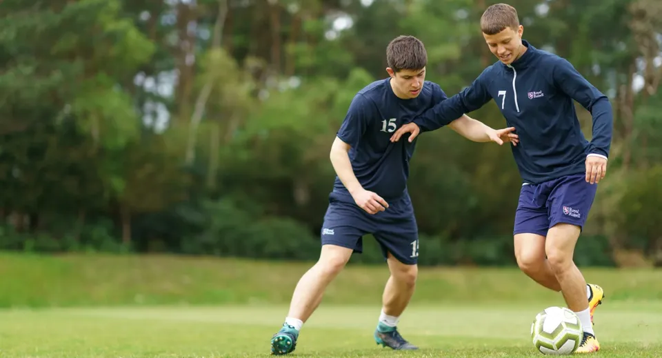 Due studenti della Royal Russell School giocano a calcio su un campo in erba circondato da alberi.