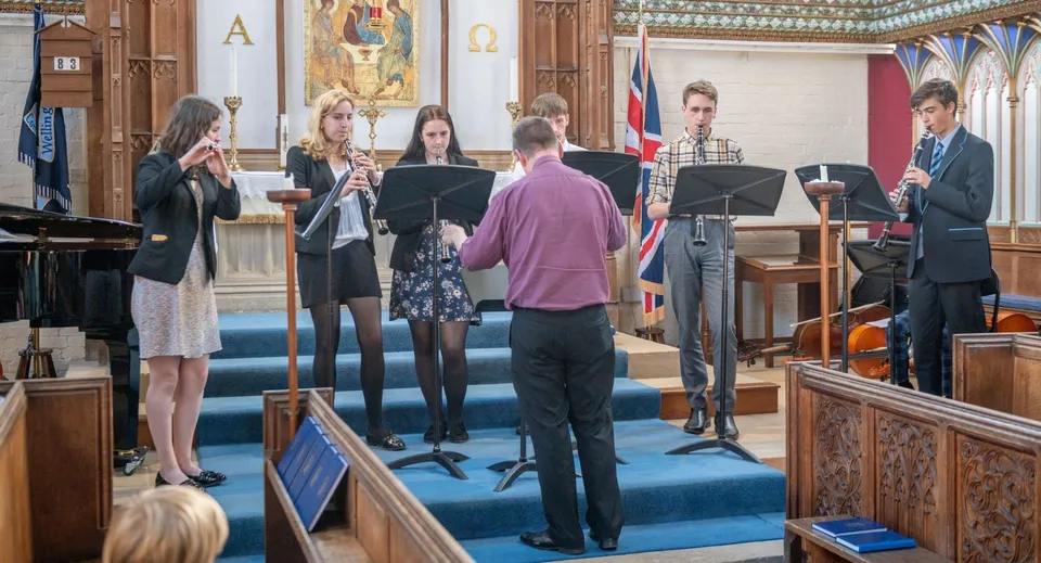 Gli studenti della Wellington School suonano strumenti a fiato in una cappella sotto la direzione di un direttore.