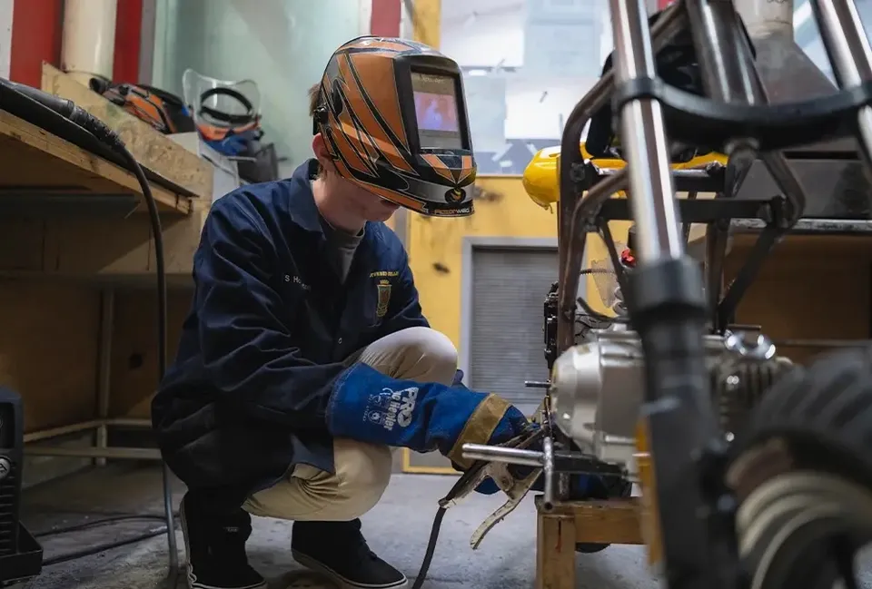 Una persona indossa una maschera da saldatore e un abbigliamento protettivo mentre lavora a un macchinario nell'officina dello Spotswood College.