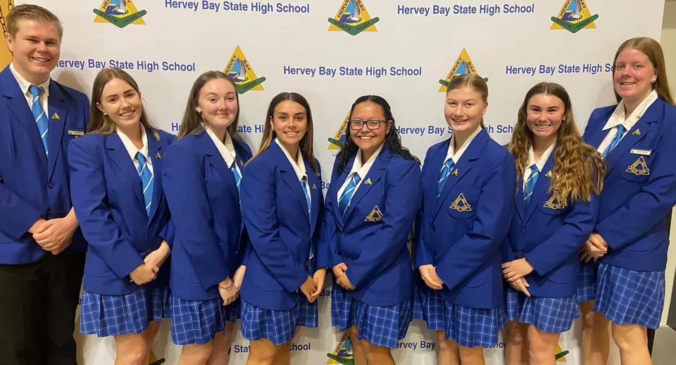 Un gruppo di studenti della Hervey Bay State High School è in posa davanti a uno sfondo con il nome e il logo della scuola.
