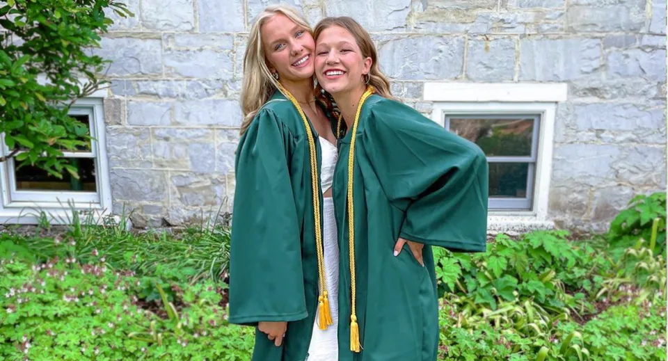Due studentesse della Burr and Burton Academy in toga verde da diploma si abbracciano davanti a un edificio in pietra circondato dal verde.