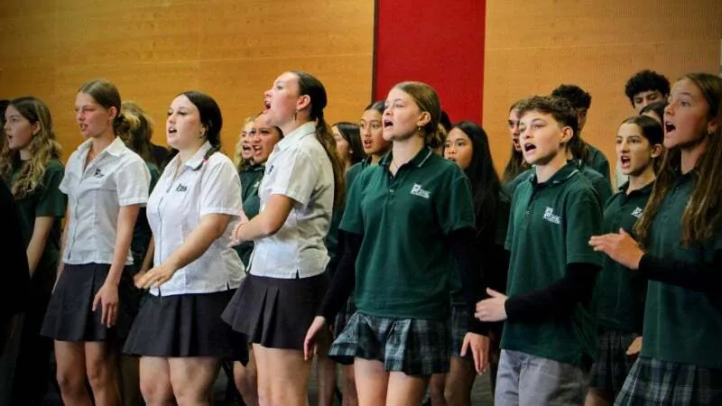 Un grupo de estudiantes de Orewa College canta junto en el escenario frente a un telón rojo.
