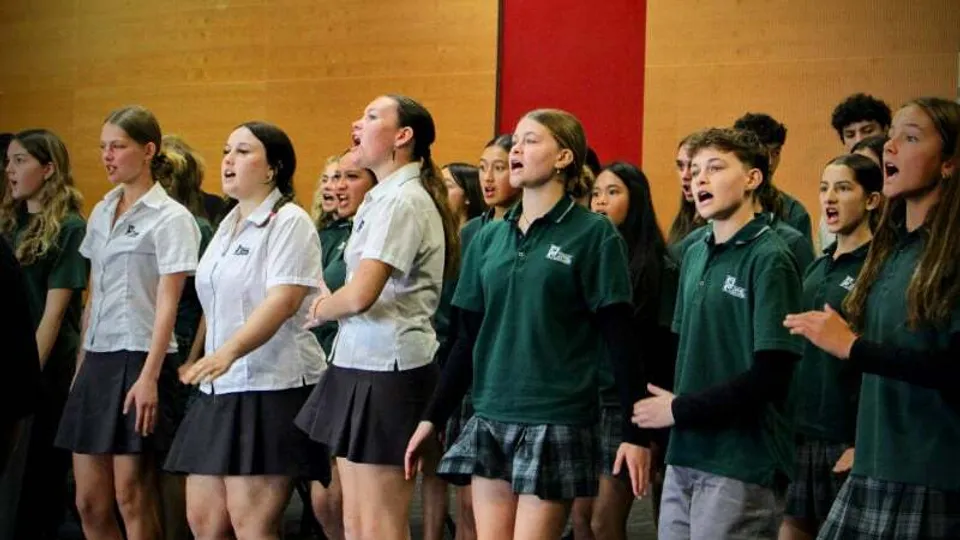 Un gruppo di studenti dell'Orewa College canta insieme sul palco davanti a un sipario rosso.