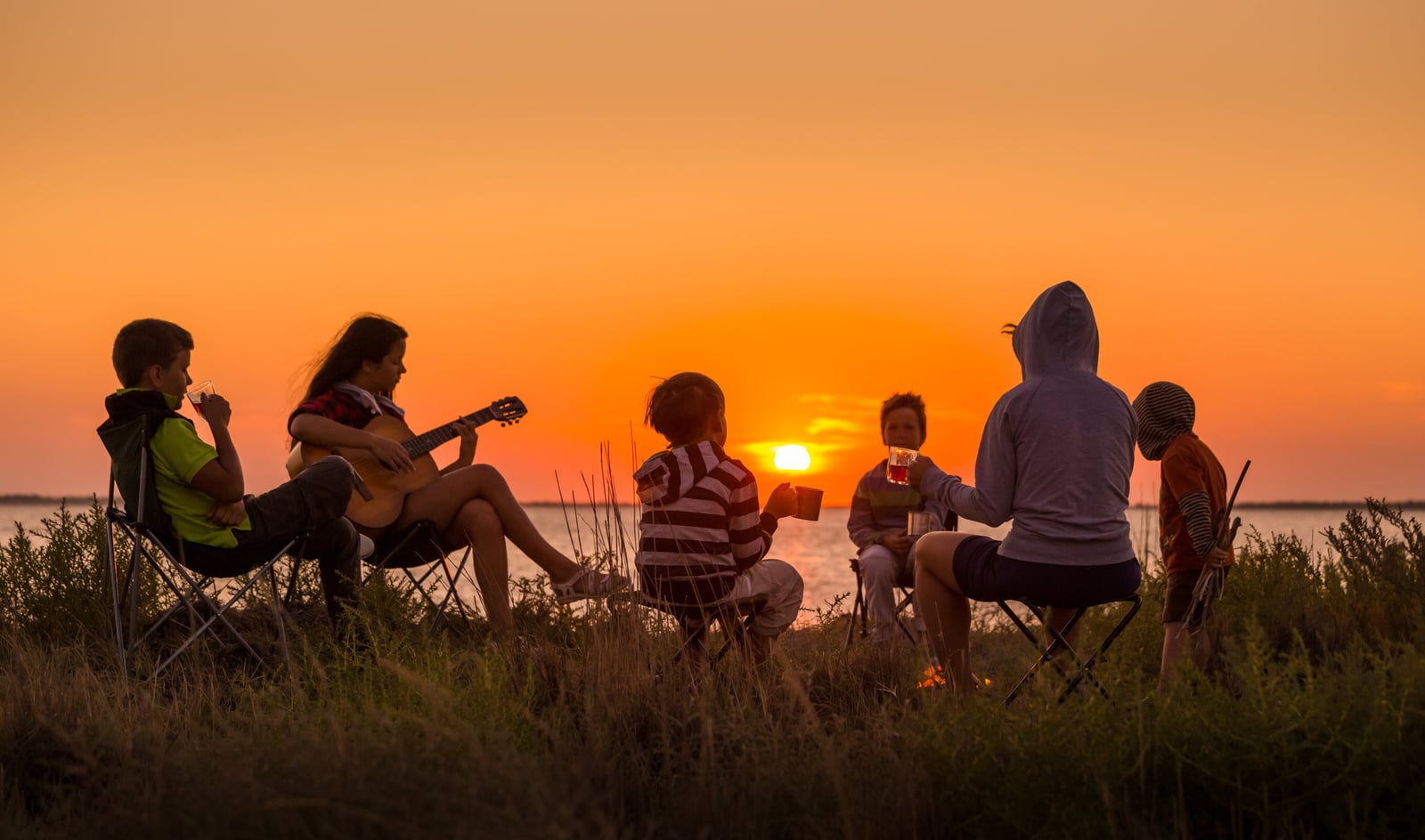 Familie sitzt bei Sonnenuntergang mit Gitarre und Getränken auf Campingstühlen am Wasser