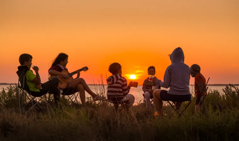 Familie sitzt bei Sonnenuntergang mit Gitarre und Getränken auf Campingstühlen am Wasser