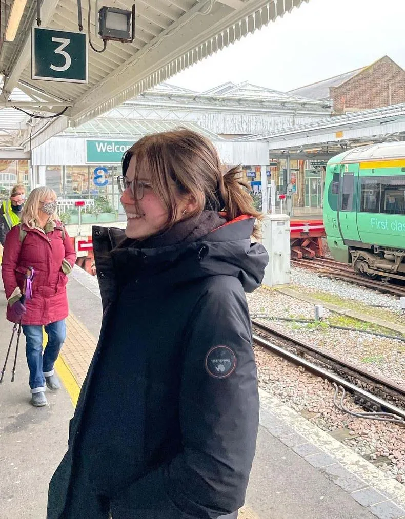 Jasmin lächelt auf einem englischen Bahnsteig, im Hintergrund ein haltender Regionalzug