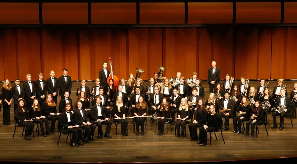 Una grande orchestra delle Jenison Public Schools si esibisce su un palco con parete posteriore in legno e file di spettatori sullo sfondo.