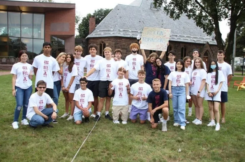 Grupo de jóvenes posando en un césped frente a un edificio escolar con camisetas blancas