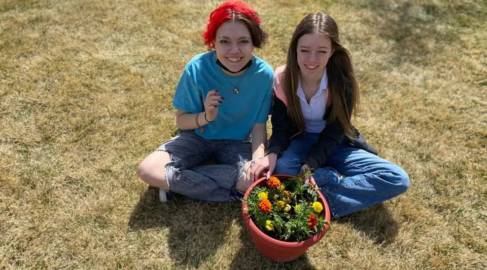 Due studentesse della Coalhurst High School siedono sull'erba tenendo un cestino colorato con delle piante.