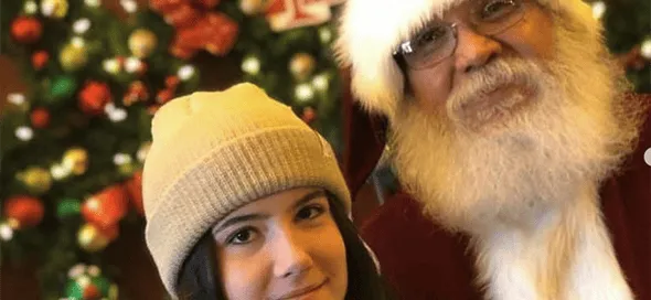 Joven con gorro beige sonríe junto a Papá Noel delante de un árbol de Navidad decorado